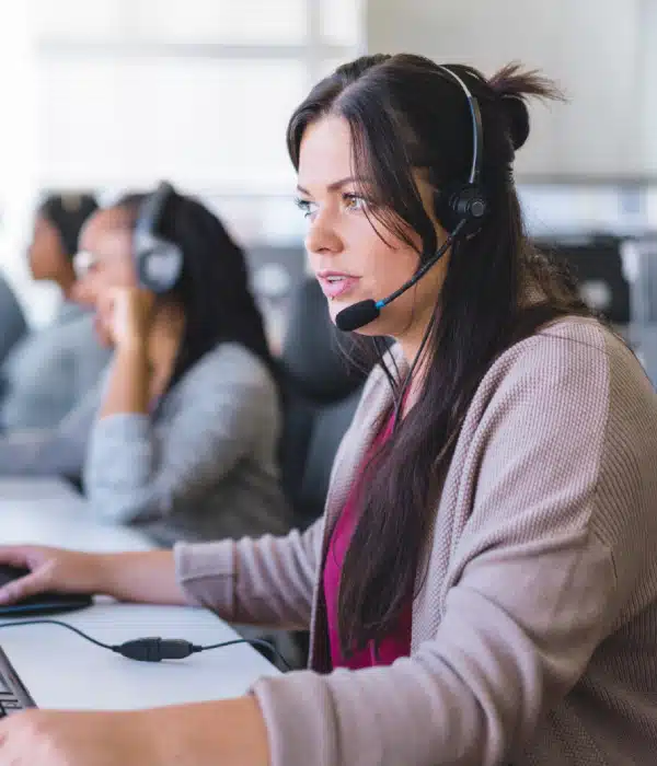 A woman wearing a headset sits at a desk, working on a computer in a call center. Other employees wearing headsets are visible in the background, focused on their screens.
