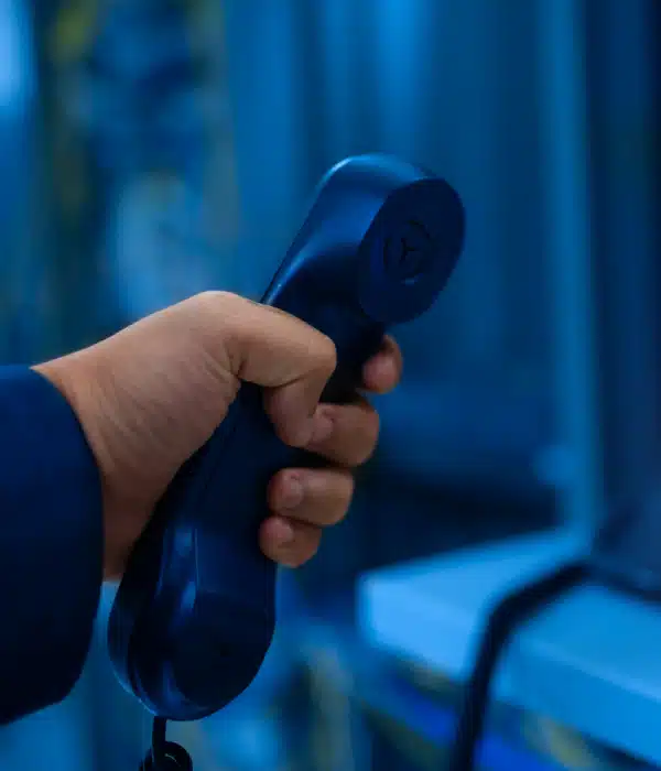 A person’s hand holding a dark blue telephone handset, with a blurred background of electronic equipment and cables, likely in a server room or data center.