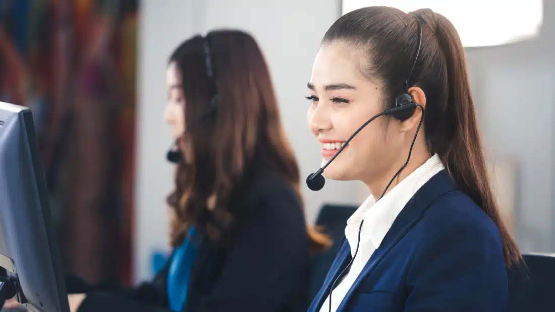 Two women wearing headsets and business attire sit at desks in an office, working on computers and smiling as they provide customer service. The focus is on the woman in the foreground.