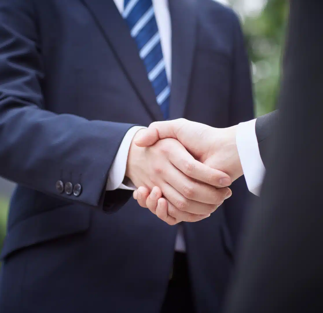 Two people wearing business suits are shaking hands outdoors, suggesting a professional agreement or partnership. Only their hands and part of their torsos are visible.
