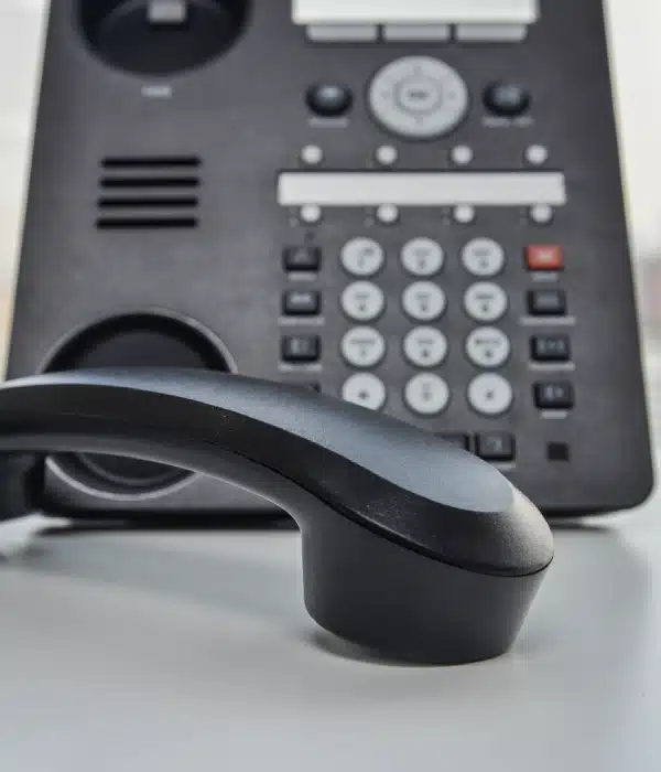 A close-up of a black office desk phone with the receiver off the hook, resting on a white surface. The phone's buttons and display screen are visible in the background.