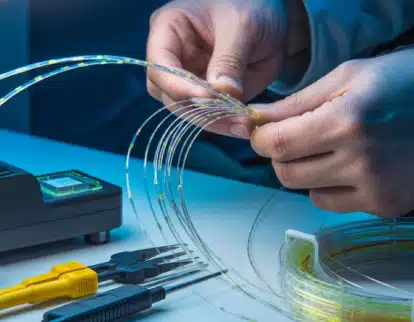 A person’s hands work with fiber optic cables at a table, surrounded by tools and equipment, preparing the cables for installation or maintenance.