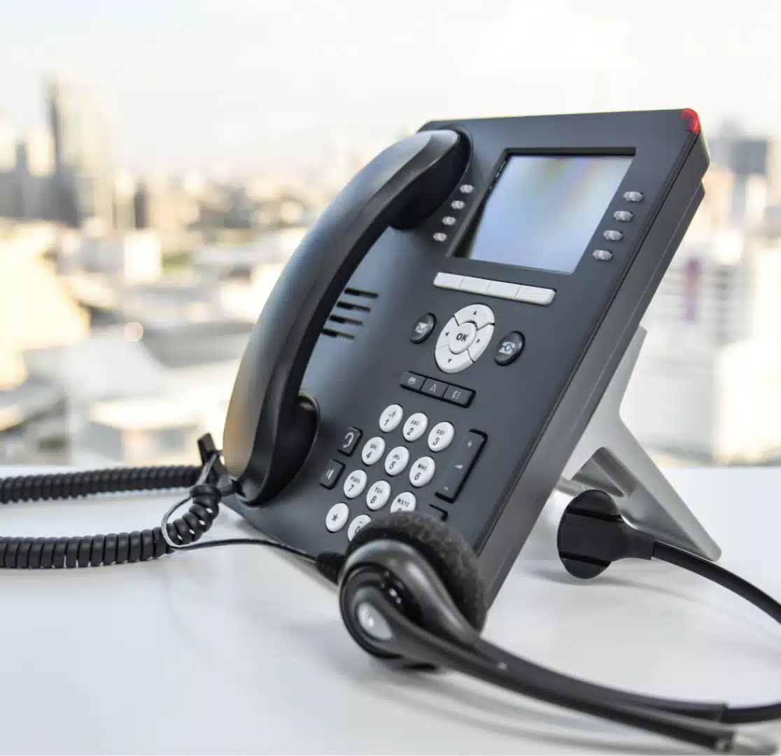 A black office desk phone with a digital display and headset rests on a white surface, with a blurred cityscape visible in the background.