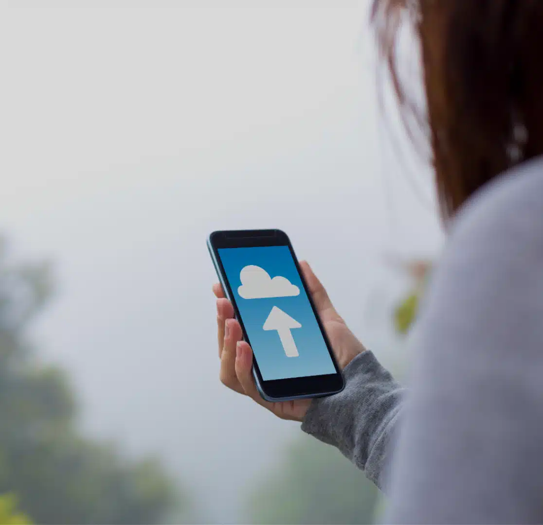 A person holds a smartphone displaying a cloud icon with an upward arrow, indicating cloud uploading, against a blurred outdoor background.
