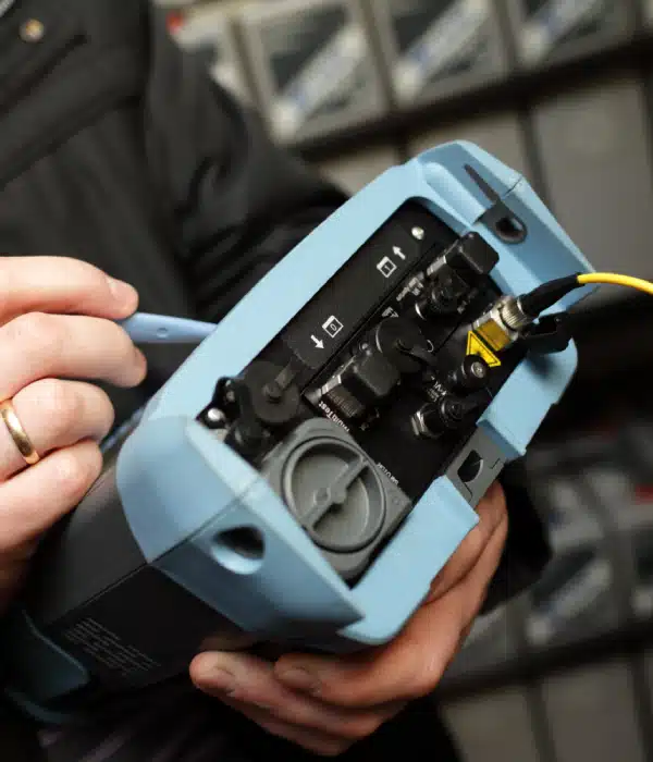 A person holds a blue handheld electronic device with various ports and cables connected, possibly using it for technical diagnostics or testing.