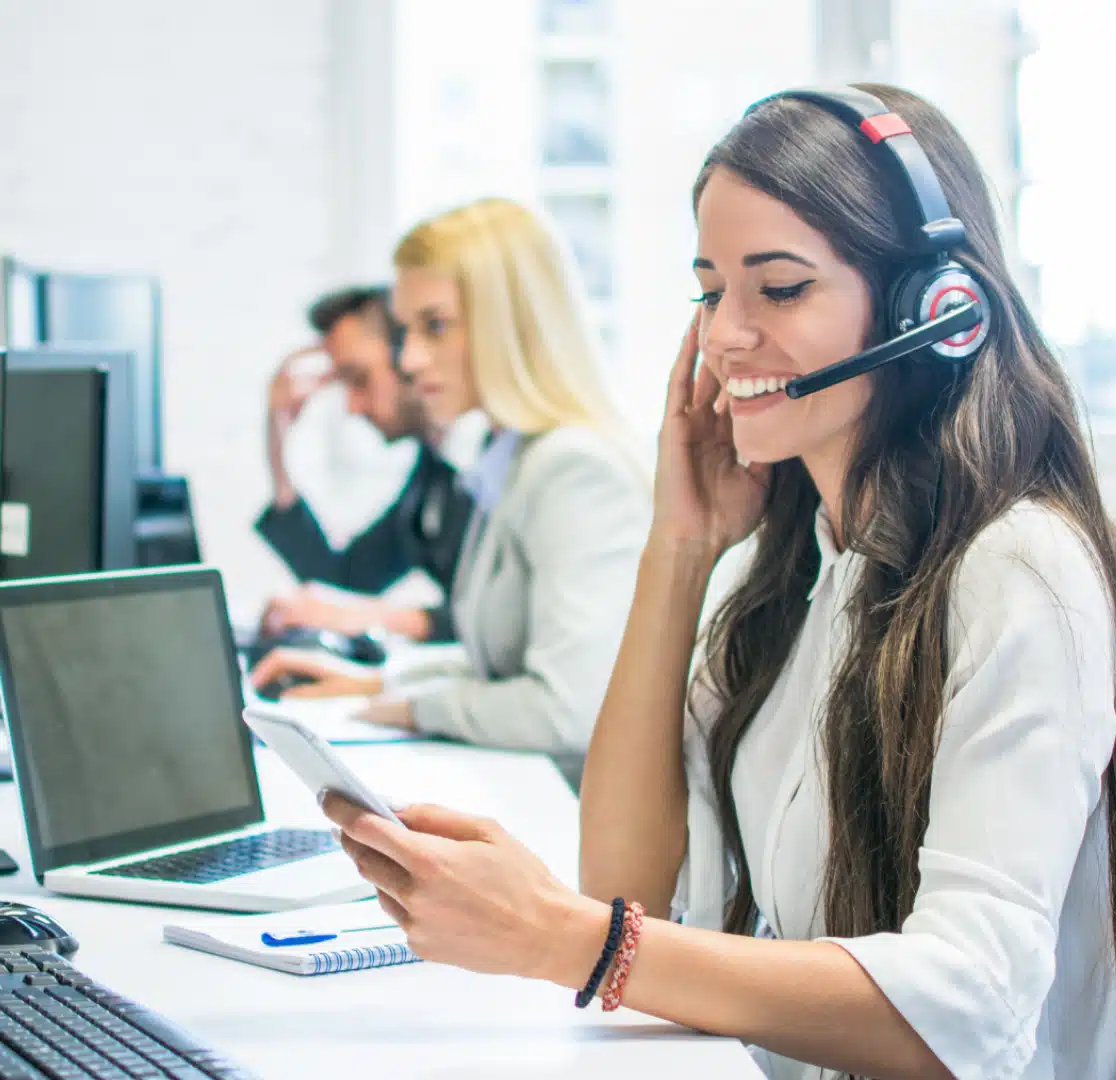 A smiling woman wearing a headset sits at a desk, holding a smartphone, with a laptop and notepad in front of her. Two other people, also wearing headsets, are working at computers in the background.